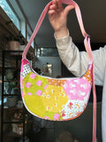 A handmade quilted crescent shoulder bag with a multicolor floral and geometric patchwork pattern, held up by a person's hand in an indoor setting.