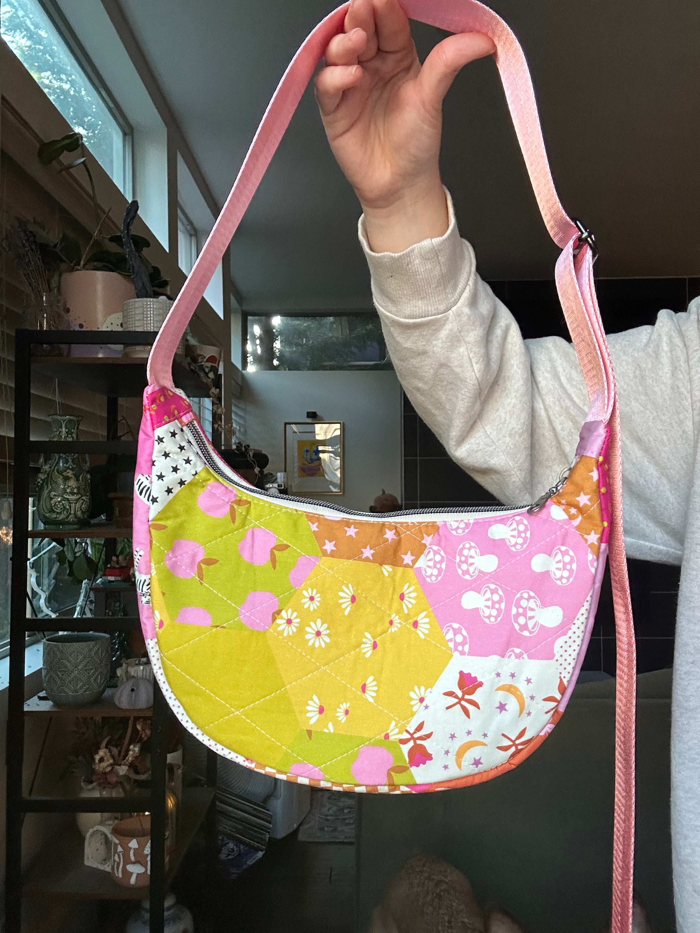 A handmade quilted crescent shoulder bag with a multicolor floral and geometric patchwork pattern, held up by a person's hand in an indoor setting.