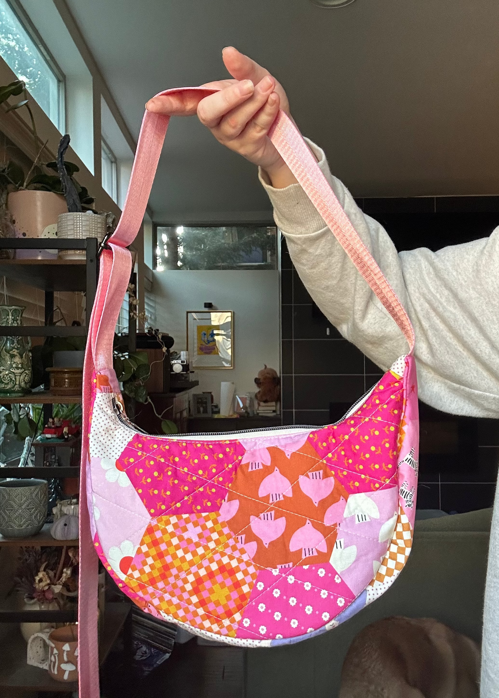 A handmade crescent-shaped shoulder bag with a pink and orange patchwork pattern, held up by a person in a home interior setting.