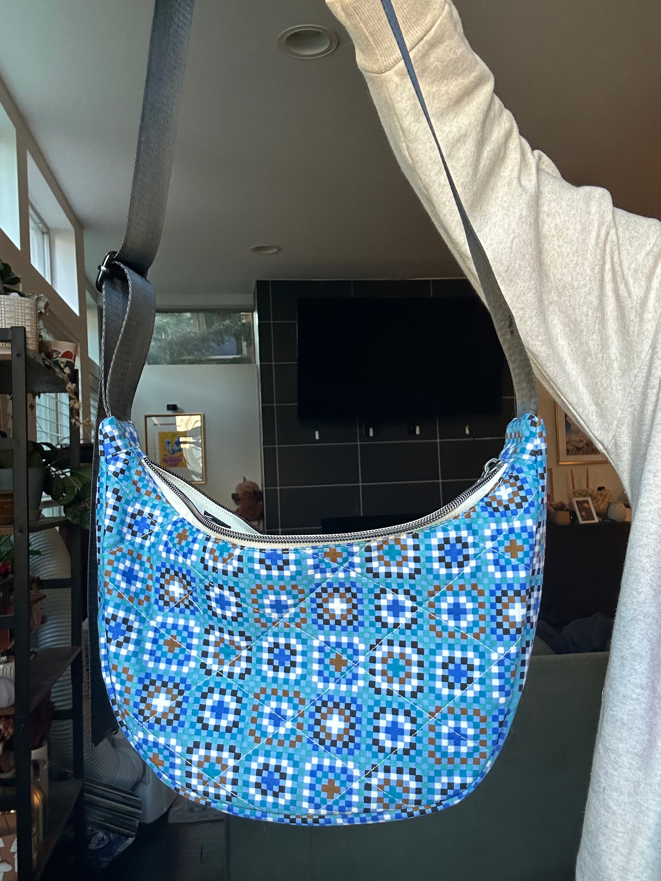 A blue patterned crescent-shaped shoulder bag with a gray strap, held up by a person in a home interior setting.