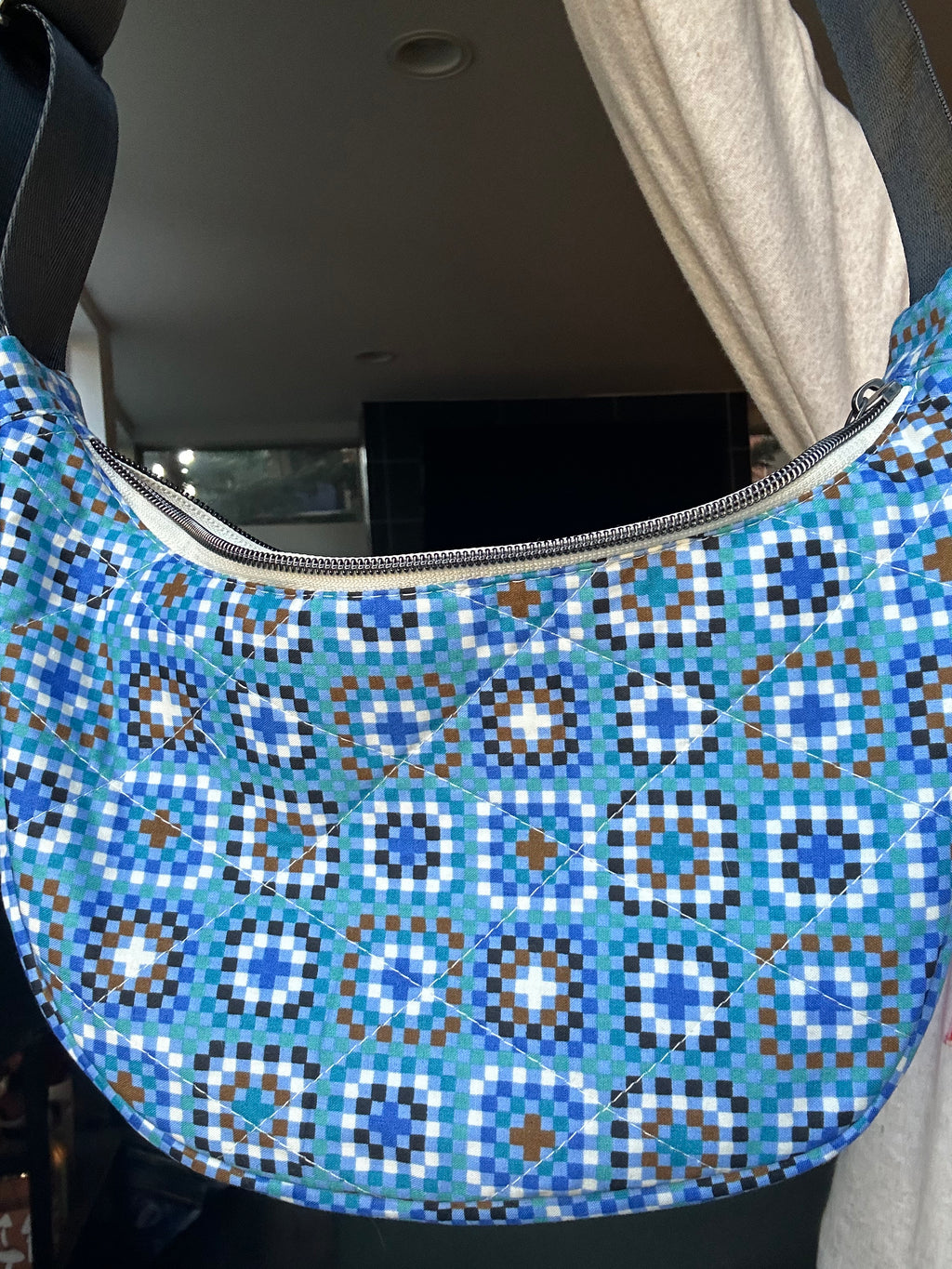 A close-up of a quilted crescent shoulder bag with a geometric medallion pattern in shades of blue, white, and brown, featuring a silver zipper closure.