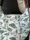 A close-up of a quilted shoulder bag featuring an all-over green duck print on an off-white background, with a brown strap and a silver zipper.