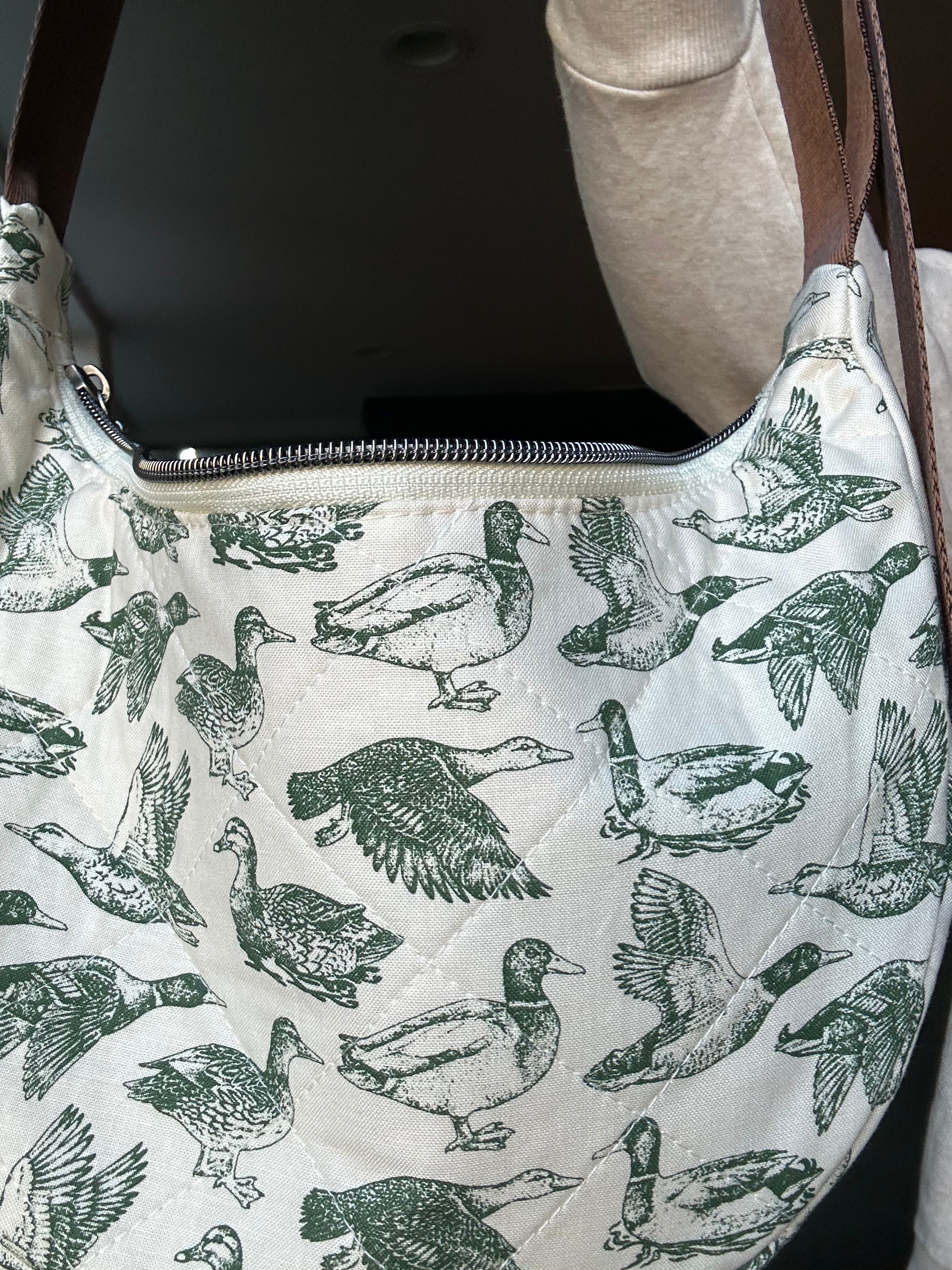 A close-up of a quilted shoulder bag featuring an all-over green duck print on an off-white background, with a brown strap and a silver zipper.