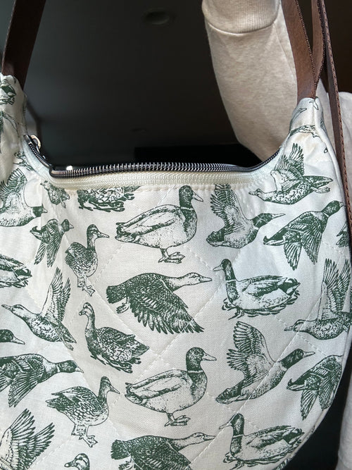 A close-up of a quilted shoulder bag featuring an all-over green duck print on an off-white background, with a brown strap and a silver zipper.
