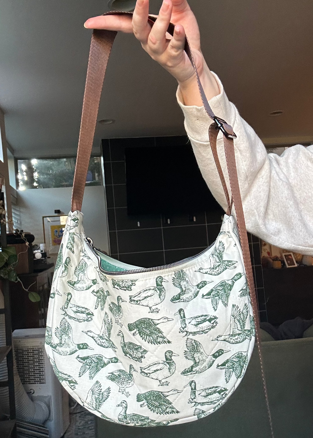 A person holds up a crescent-shaped shoulder bag with a brown strap. The bag has a white base with a green pattern of birds and leaves, and is photographed in a home interior setting.