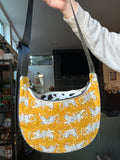 A quilted crescent-shaped shoulder bag with a yellow and white tiger print, held up by a person's hand in a living room setting.