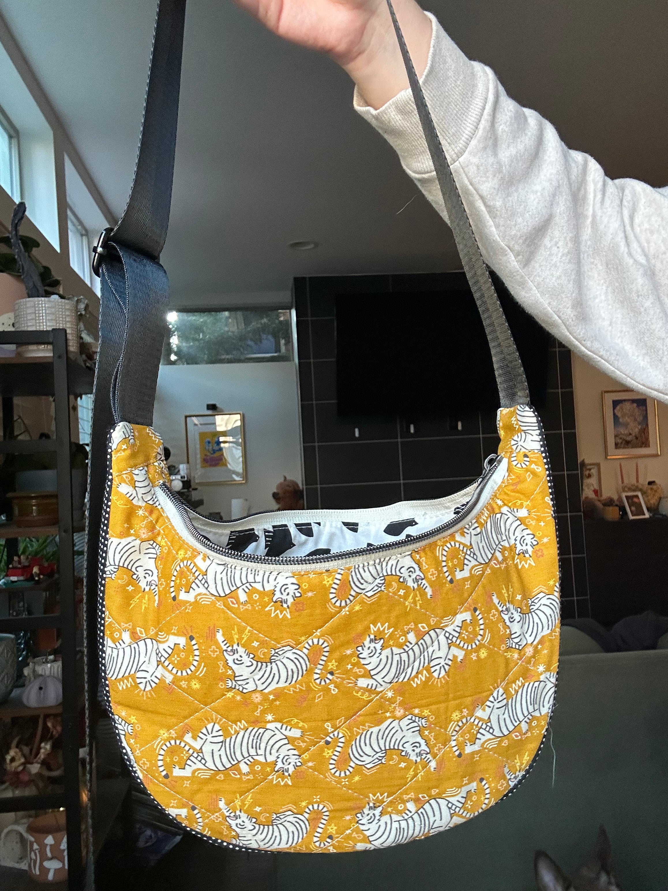 A quilted crescent-shaped shoulder bag with a yellow and white tiger print, held up by a person's hand in a living room setting.