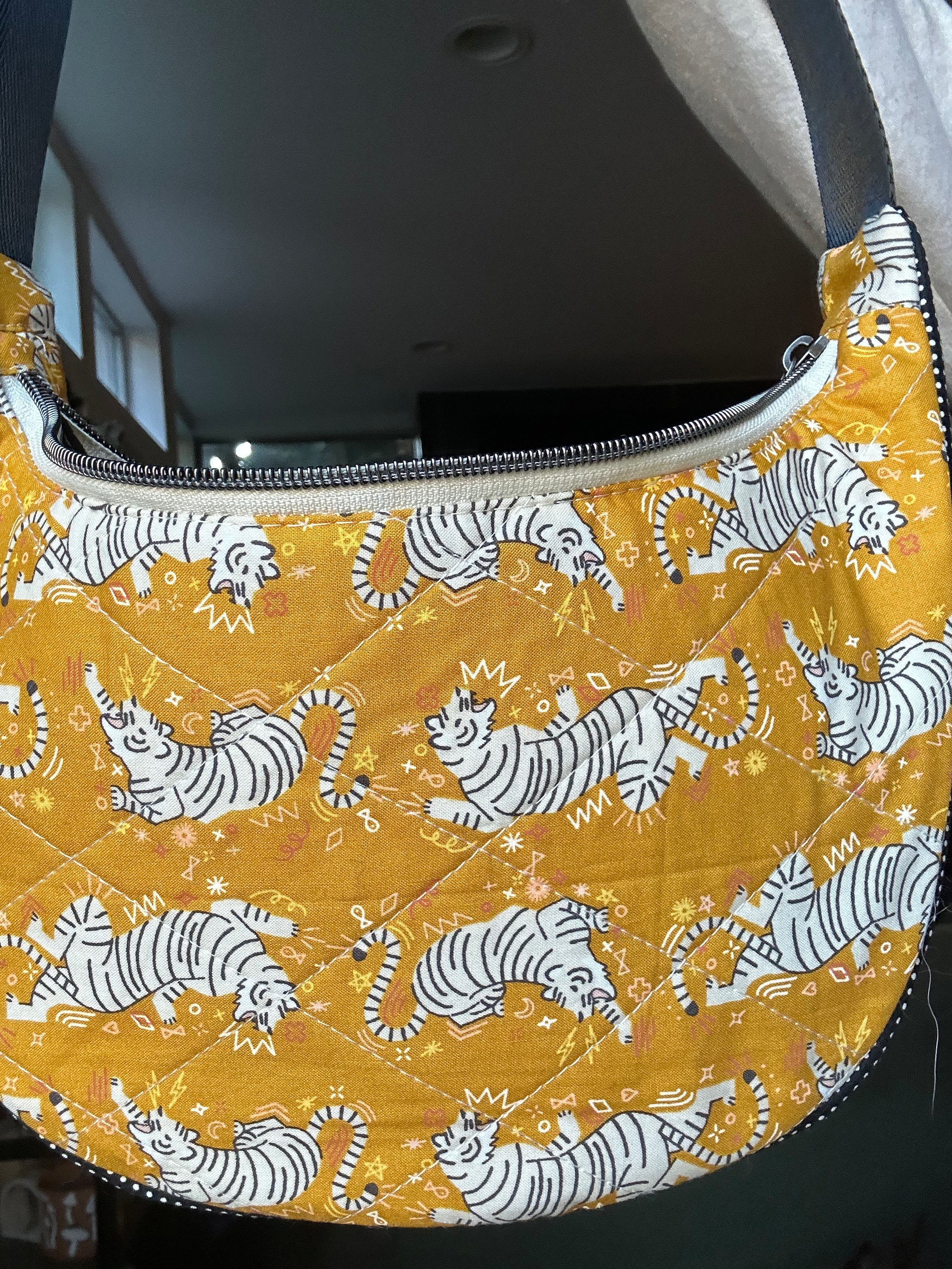 A quilted crescent-shaped shoulder bag with a mustard yellow background and an all-over print of white tigers with small gold crowns. The bag features a silver zipper closure and a dark strap, photographed in an indoor setting.