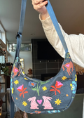 A person holds up a crescent-shaped quilted shoulder bag with a blue-gray base and a colorful print of pink rabbits, flowers, and stars. The bag has a blue strap and is shown in a home interior setting.