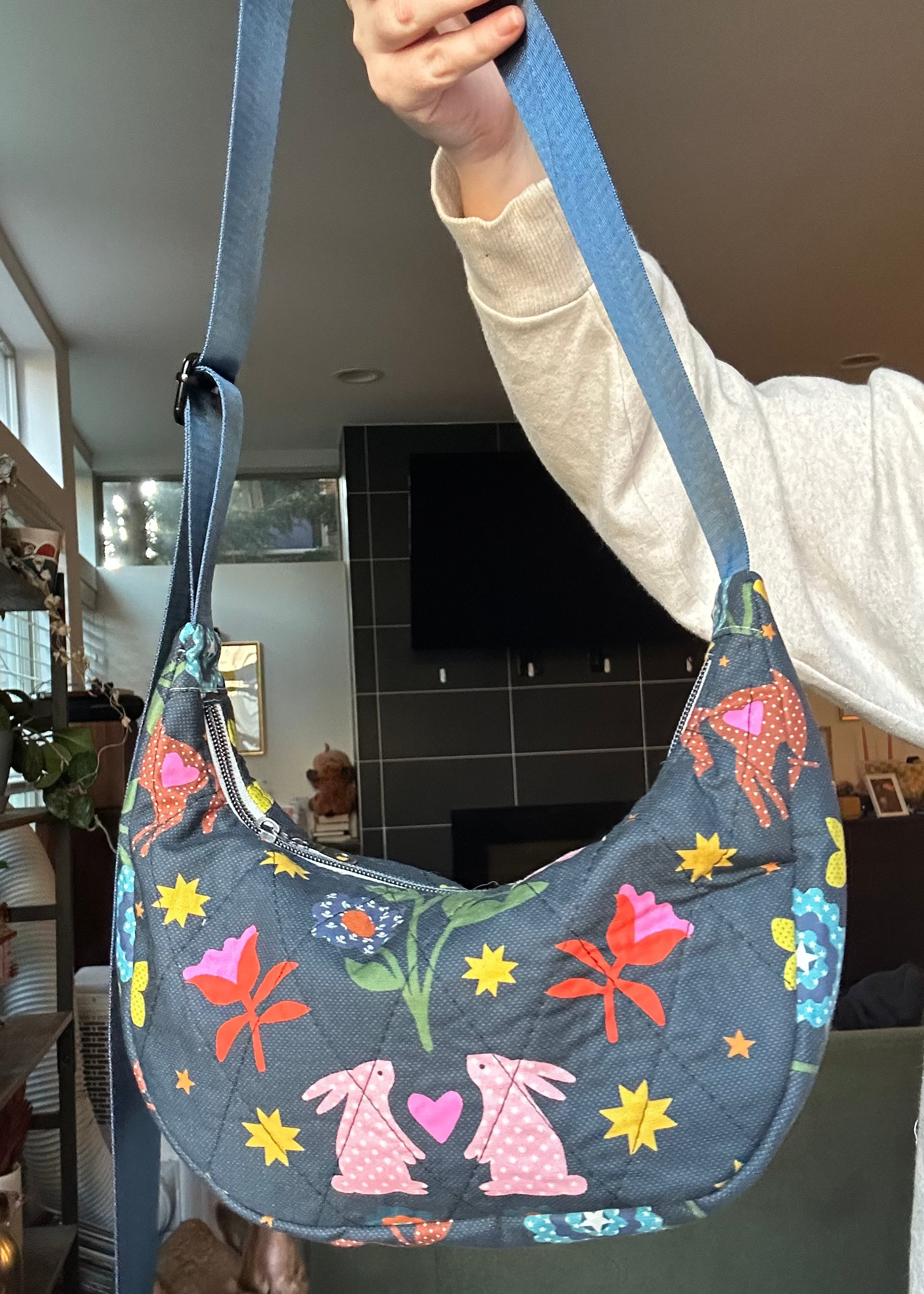 A person holds up a crescent-shaped quilted shoulder bag with a blue-gray base and a colorful print of pink rabbits, flowers, and stars. The bag has a blue strap and is shown in a home interior setting.