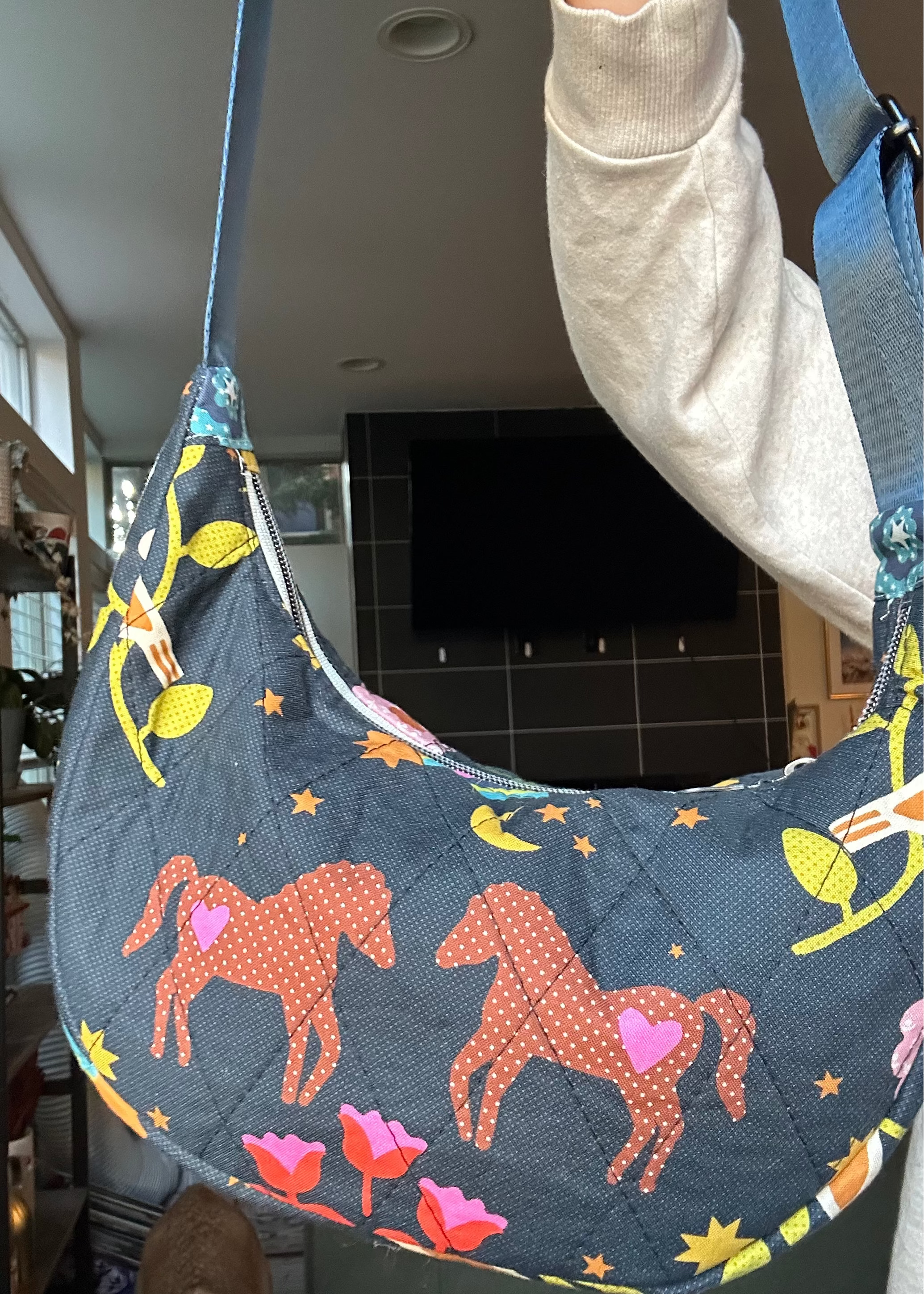A person holds up a crescent-shaped shoulder bag with a dark slate blue background and a whimsical print of orange horses, pink tulips, yellow stars, and green leaves. The bag has a quilted texture and is shown in an indoor home environment.