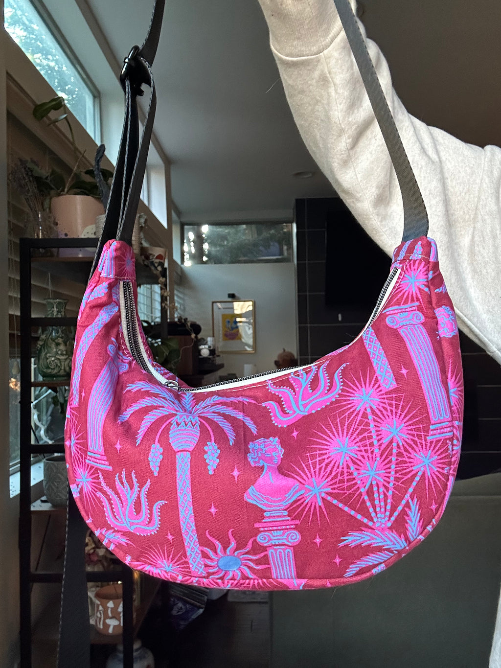 A pink crescent-shaped shoulder bag with a light blue palm tree and classical bust pattern, held up by a person wearing a white sweatshirt in an indoor setting.
