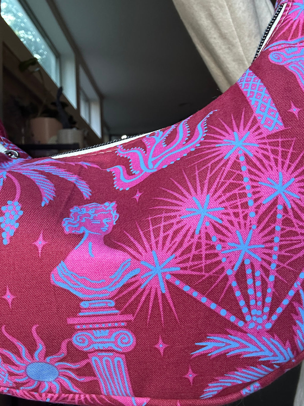 A close-up of a pink and blue patterned fabric item, likely a bag or pouch, featuring classical busts, stars, and palm-like motifs on a magenta background.