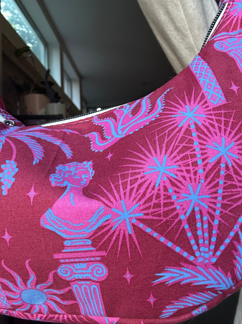 A close-up of a pink and blue patterned fabric item, likely a bag or pouch, featuring classical busts, stars, and palm-like motifs on a magenta background.