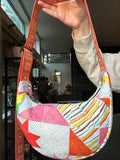 A handmade crescent-shaped shoulder bag with a colorful geometric patchwork pattern, held up by a person's hand in an indoor setting.