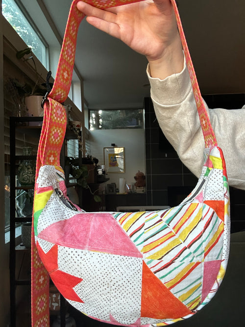 A handmade crescent-shaped shoulder bag with a colorful geometric patchwork pattern, held up by a person's hand in an indoor setting.