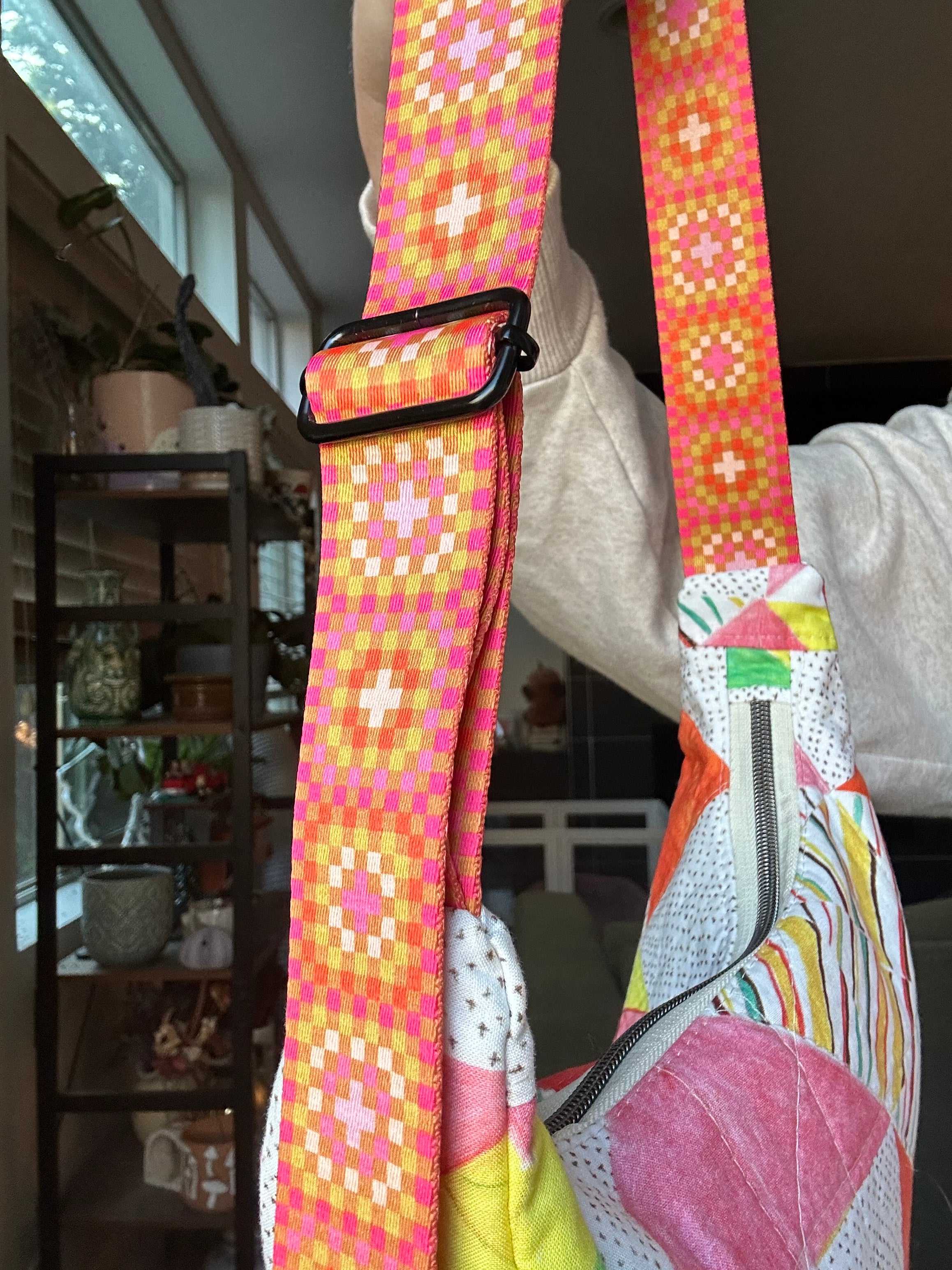 A close-up of a colorful crossbody bag strap with a pink, orange, and yellow geometric pattern, worn by a person in an indoor home setting.