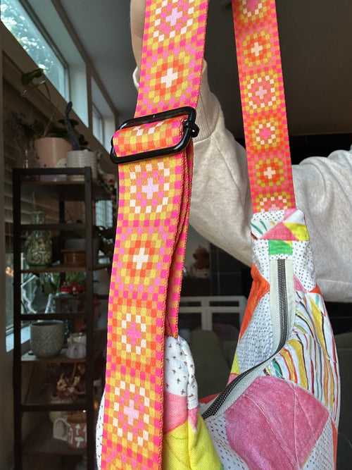 A close-up of a colorful crossbody bag strap with a pink, orange, and yellow geometric pattern, worn by a person in an indoor home setting.
