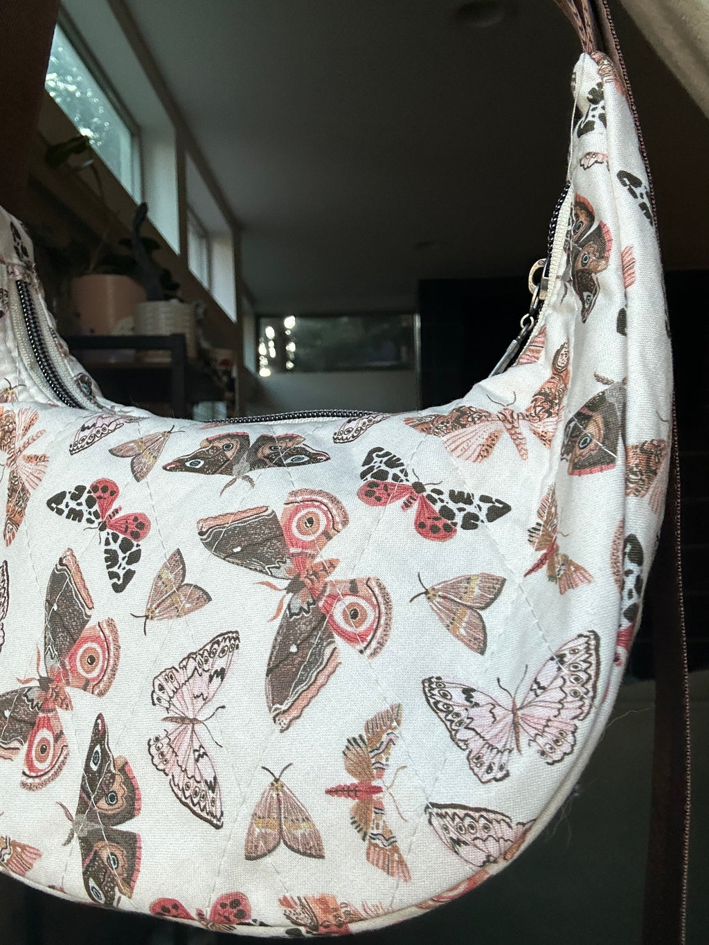 A crescent-shaped shoulder bag with an all-over butterfly and moth print on a white background, photographed in a real indoor environment.