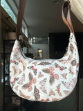 A crescent-shaped shoulder bag with a white background and an all-over butterfly and moth print in muted pink, brown, and gray tones, featuring a brown shoulder strap.