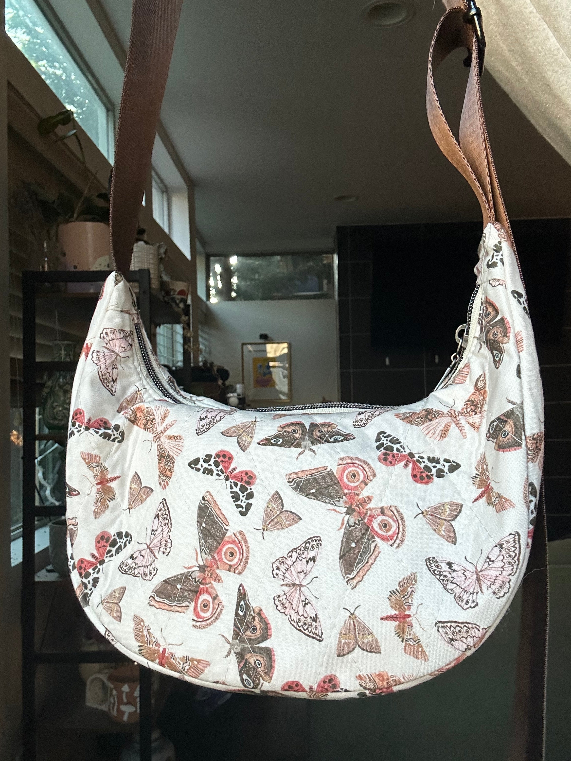 A crescent-shaped shoulder bag with a white background and an all-over butterfly and moth print in muted pink, brown, and gray tones, featuring a brown shoulder strap.