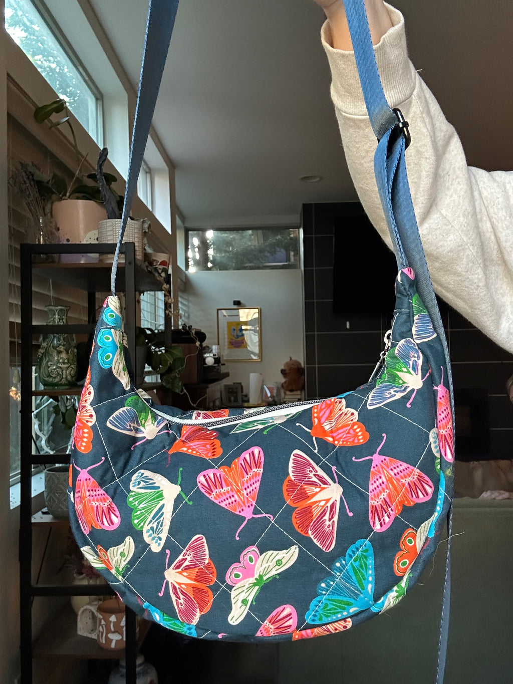 A person holds up a crescent-shaped shoulder bag with a dark blue background and a colorful butterfly print. The bag has a blue strap and is shown in a home interior setting.