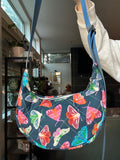 A person holds up a crescent-shaped shoulder bag with a dark blue background and a colorful butterfly print. The bag has a blue strap and is shown in a home interior setting.