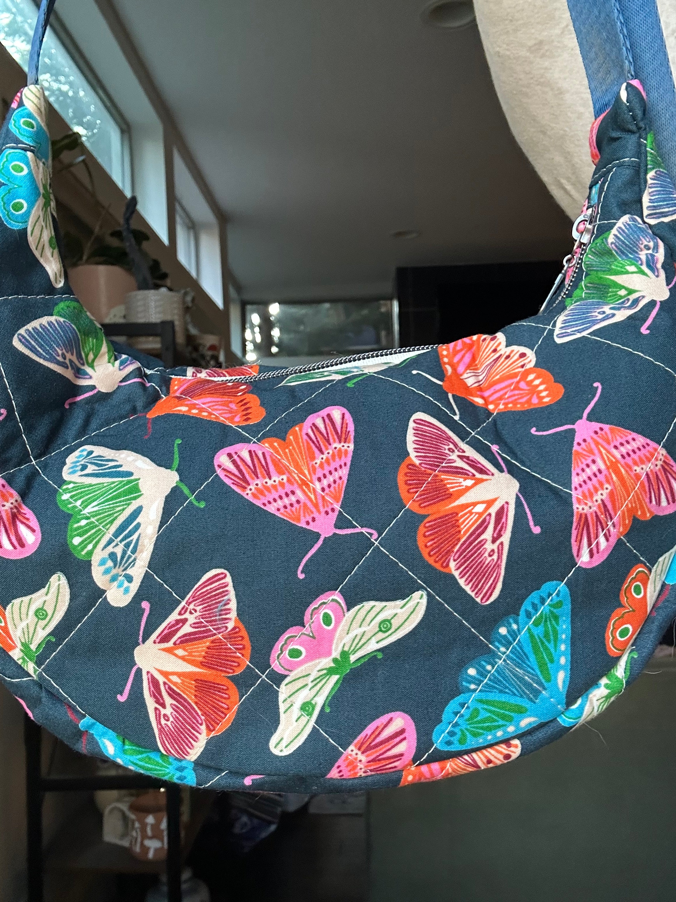 A crescent-shaped shoulder bag with a dark navy blue background and a colorful butterfly print, shown hanging indoors.